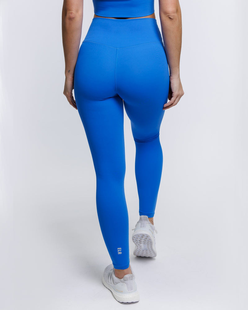 A female model showing the back of the ButterBod Hourglass Leggings in electric blue colour. The high-waisted wrap design and V-cut waistband accentuate her curves, while the booty-lifting back seam provides a flattering lift. The leggings have a soft matte finish, medium compression, and are made from 4-way stretch fabric for maximum comfort and versatility.