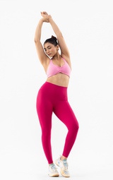 A woman in a pink sports bra and magenta high-waisted leggings poses in a side stretch against a solid white background.