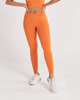 A female model wearing a orange Hourglass Leggings. The high-waisted wrap design and V-cut waistband accentuate her curves, while the booty-lifting back seam provides a flattering lift. The legging has a soft matte finish, medium compression, and is made from 4-way stretch fabric for maximum comfort and versatility