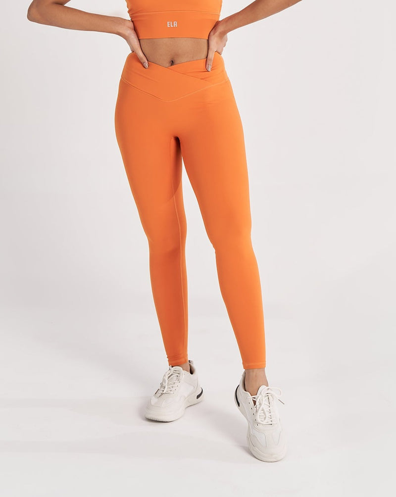 A female model wearing a orange Hourglass Leggings. The high-waisted wrap design and V-cut waistband accentuate her curves, while the booty-lifting back seam provides a flattering lift. The legging has a soft matte finish, medium compression, and is made from 4-way stretch fabric for maximum comfort and versatility