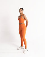 An athletic female model wearing the ButterBod Hourglass Leggings in orange and matching halter neck sports bra, highlighting it as a coordinated set. The high-waisted criss cross design accentuate her curves, while the booty-lifting back seam provides a flattering lift. The fabric has a soft finish, medium compression, and are made from 4-way stretch fabric for maximum comfort and versatility making it best choice for gym and yoga