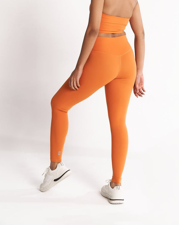 A female model showing the back of the ButterBod Hourglass Leggings in orange colour. The high-waisted wrap design and V-cut waistband accentuate her curves, while the booty-lifting back seam provides a flattering lift. The leggings have a soft matte finish, medium compression, and are made from 4-way stretch fabric for maximum comfort and versatility.