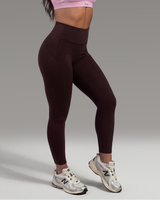Woman wearing a pink sports bra and dark brown leggings on a gray background