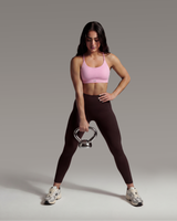 Woman in pink sports bra and black leggings holding a silver kettlebell against a gray background