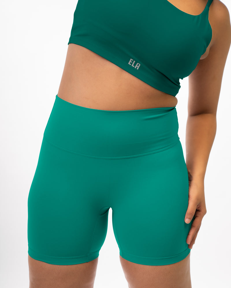 plus size Female model posing in premium green high waisted and squat proof bike shorts suitable for athletic activities with dark green sports bra giving a monochromatic athleisure look