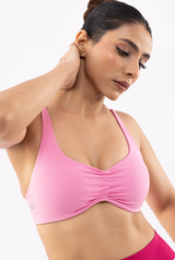 A woman wearing a light pink sports bra with a gathered front and wide straps, shown in a close-up studio shot against a plain white background.