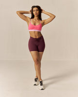 Woman wearing a two tone pink sports bra and maroon brown shorts on a beige background