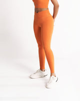 A female model wearing a orange Hourglass Leggings. The high-waisted wrap design and V-cut waistband accentuate her curves, while the booty-lifting back seam provides a flattering lift. The legging has a soft matte finish, medium compression, and is made from 4-way stretch fabric for maximum comfort and versatility