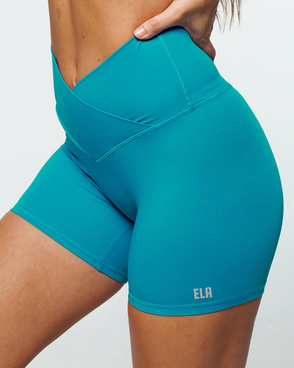 Close up of Turquoise Bike shorts on a female fit model highlighting Pinch-free Criss Cross High Waistband, Anti-Camel Toe, Seam-Free Front Panel, Slimming + Buttery-Smooth, and Squat-Proof features