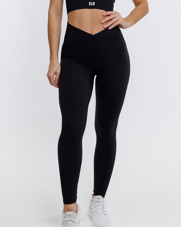 A female model wearing a black Hourglass Leggings, highlighting her hourglass figure. The high-waisted wrap design and V-cut waistband accentuate her curves, while the booty-lifting back seam provides a flattering lift. The leggings have a soft matte finish, medium compression, and are made from 4-way stretch fabric for maximum comfort and versatility