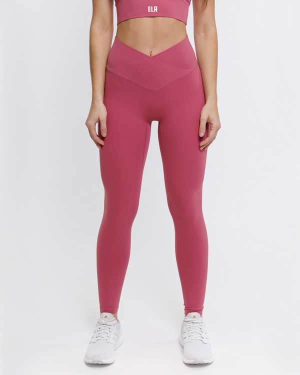 A female model wearing a pink Hourglass Leggings, highlighting her hourglass figure. The high-waisted wrap design and V-cut waistband accentuate her curves, while the booty-lifting back seam provides a flattering lift. The leggings have a soft matte finish, medium compression, and are made from 4-way stretch fabric for maximum comfort and versatility