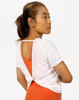 Female model is showing the White gym tee that has a round scoop neck in the front made of fine mesh fabric with a flattering V-shaped twisted open back design. The top is perfect for use as a cover-up on top of a sports bra and is great for gym, yoga, or everyday wear. 