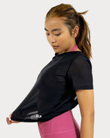 Female model is showing the black gym tee that has a round scoop neck in the front made of fine mesh fabric with a flattering V-shaped twisted open back design. The top is perfect for use as a cover-up on top of a sports bra and is great for gym, yoga, or everyday wear. 